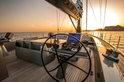 A large yacht's helm and seating area are in the foreground, with a warm sunset over the ocean and distant hills in the background.