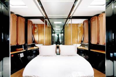 A white-sheeted bed with two pillows sits in the foreground of a luxurious, mirrored room with wooden cabinets lining the walls and a bright ceiling.