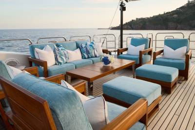 A luxurious outdoor seating area with light blue cushions and wooden frames is set on a yacht's deck, with the calm ocean and a distant, tree-covered coastline in the background.
