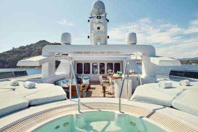 A luxurious yacht's jacuzzi and sun loungers in the foreground are set against a backdrop of the open sea and a distant, tree-covered island.