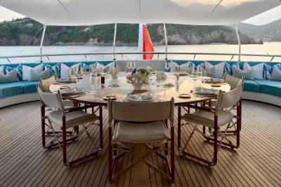A beautifully set dining table on a yacht's deck is surrounded by plush seating, with a scenic coastline visible in the background.