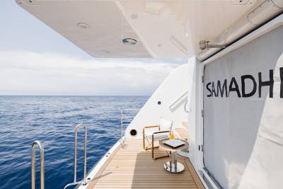 A wooden deck with chairs and a drink sits on the stern of a yacht, with the blue ocean and sky stretching out in the background.