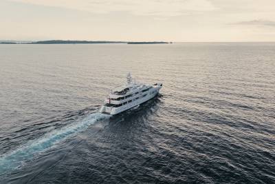 A large white yacht cruises through calm, dark water, leaving a foamy wake, with a distant, hazy coastline under a pale sky.