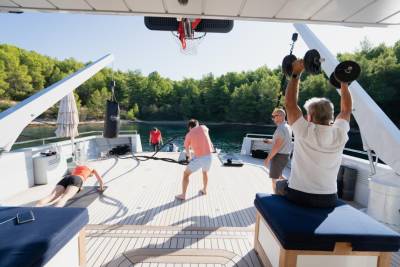 People exercise on the deck of a yacht with a lush green coastline in the background.