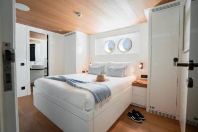 A neatly made bed with blue and white striped pillows and a straw hat sits in the foreground of a bright, modern cabin with porthole windows in the background.