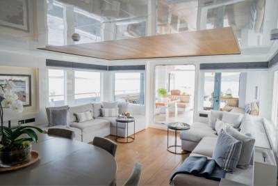 A spacious, modern living area with a large sectional sofa and dining table in the foreground, and a bright, open space with large windows overlooking a body of water in the background.