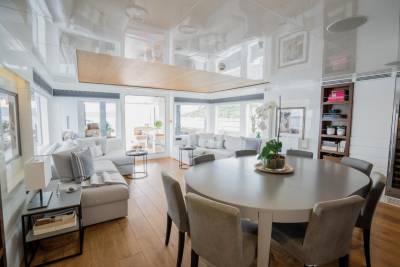 A round dining table with upholstered chairs sits in the foreground, while a spacious living area with a sectional sofa and large windows fills the background.