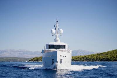A white yacht cuts through blue water in the foreground, with a tree-covered island and hazy mountains in the background under a clear sky.