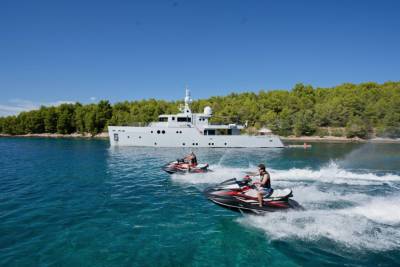 Two jet skis race across the turquoise water in the foreground, with a large yacht anchored in the background near a tree-covered island.