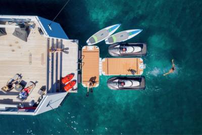 People relax on a yacht's deck and swim in the clear blue ocean alongside jet skis and paddleboards.