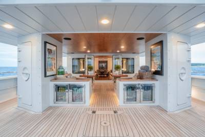 A spacious, well-lit deck with wooden flooring in the foreground leads to a bar area with refrigerators and artwork, opening into a dining area with mirrors and tables in the background.