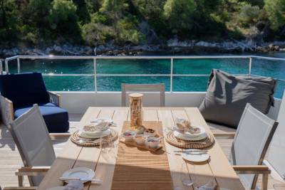 A dining table set for a meal on a yacht's deck overlooks a tranquil turquoise sea and lush green hills.