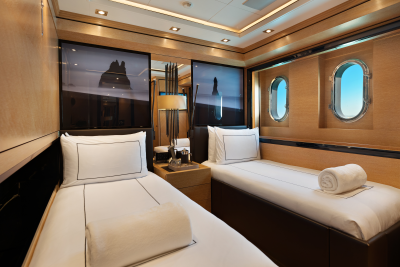 Two neatly made twin beds with rolled towels occupy the foreground, while oval windows revealing a blue sky and wooden-paneled walls form the background of this luxurious cabin.