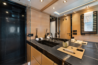 A modern bathroom with a black marble countertop and sink in the foreground, and a mirrored wall reflecting the shower and wooden accents in the background.