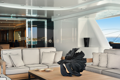 A modern yacht's outdoor lounge area with plush white sofas and a striking black sculpture in the foreground, opens to a bright blue ocean and sky in the background.