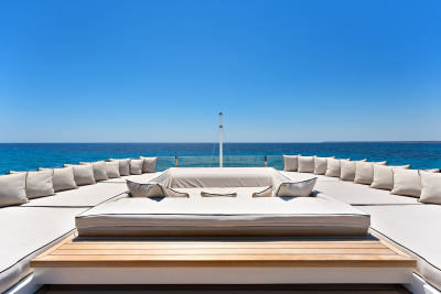 Plush white cushions arranged on a wooden deck face a vast expanse of blue ocean under a clear sky.