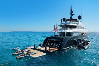 A luxurious yacht with jet skis docked at its stern floats on a calm, blue sea under a clear sky.