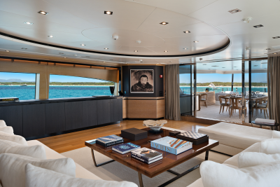 A luxurious yacht interior with a plush white sofa and coffee table in the foreground, and a scenic ocean view with an outdoor dining area in the background.