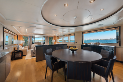 A circular dining table with chairs in the foreground is set against a backdrop of a luxurious living area and a panoramic view of the ocean through large windows.