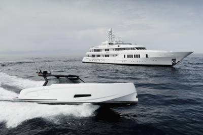 A sleek, modern white speedboat cuts through dark blue water in the foreground, with a large, luxurious white yacht sailing in the hazy background.