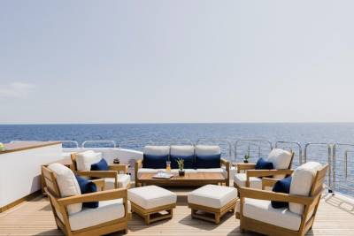 A luxurious outdoor seating area with white cushions and navy accents is arranged on a wooden deck, with the calm blue ocean and clear sky stretching out in the background.