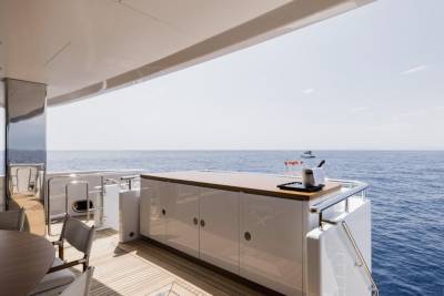 A modern yacht deck with a bar and seating in the foreground overlooks a calm blue ocean with a distant boat in the background.