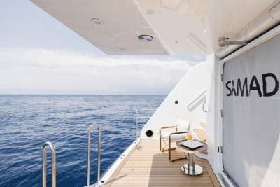 A wooden deck with two chairs and a table sits on the stern of a yacht, with the calm blue ocean stretching to the horizon under a cloudy sky.