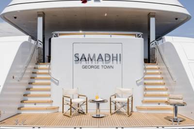 Two white chairs and a small table with drinks sit on the wooden deck of a large white yacht named "Samadhi," with a clear blue sky in the background.