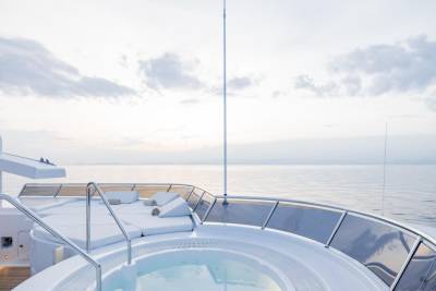 A circular jacuzzi and white lounge seating are in the foreground of a yacht, with a calm ocean and cloudy sky in the background.