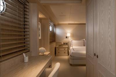 A light wood desk with a chair in the foreground leads to a neatly made bed in the background, all within a well-lit, modern interior.