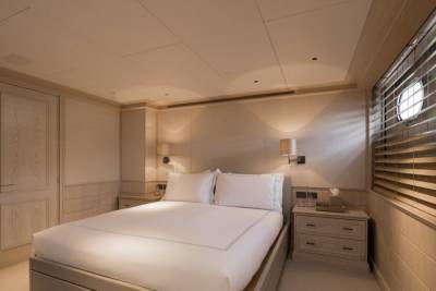 A neatly made bed with white linens dominates the foreground, while the background features a closed door and porthole windows with blinds.