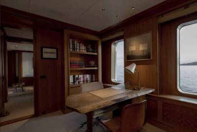 A rustic wooden desk with a leather chair sits in front of a bookshelf filled with books, while large windows reveal a calm ocean view.