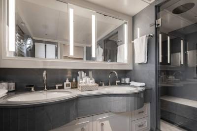 A modern, gray bathroom with a double vanity and toiletries in the foreground, and a glass-enclosed shower with towels in the background.
