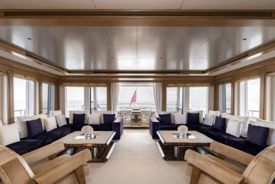 Plush navy sofas with white pillows flank ornate coffee tables in the foreground, while the background reveals a sunlit deck with a red flag visible through large windows.