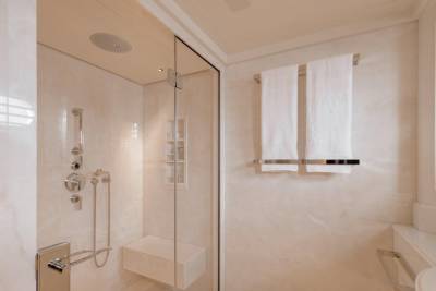 A modern, white marble shower stall with a built-in bench is in the foreground, while two fluffy white towels hang on a rack against a marble wall in the background.