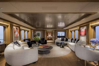 Plush white sofas and armchairs surround a circular rug in the foreground, while large windows offer views of the ocean in the background.