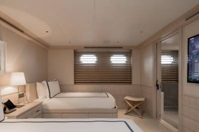 Two neatly made beds with white linens and blue trim are in the foreground, while the background features a large television screen and two circular windows with blinds.