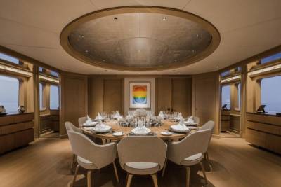 A circular dining table set for a meal is in the foreground, while a colorful abstract painting hangs on the wall in the background, framed by large windows overlooking the ocean.
