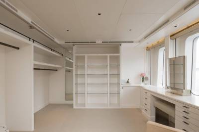 A spacious, well-lit walk-in closet with ample shelving and hanging space is situated in the foreground, while large windows offering a view of the water dominate the background.