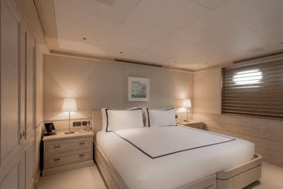 A luxurious yacht bedroom features a neatly made bed in the foreground and porthole windows with blinds in the background.