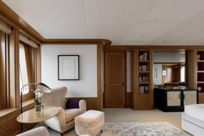 A plush armchair and ottoman sit in the foreground of a well-lit room with large, curved windows, while a built-in bookshelf and bar area are visible in the background.