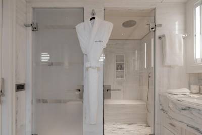 A plush white bathrobe hangs on a glass door in the foreground, while a modern, marble-clad shower with a rainfall showerhead is visible in the background.