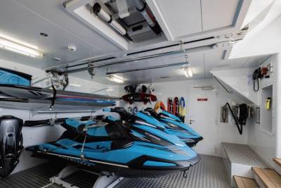 Two bright blue jet skis are parked in the foreground of a storage room filled with water sports equipment like surfboards and paddles, all neatly organized against a white, industrial-looking background.