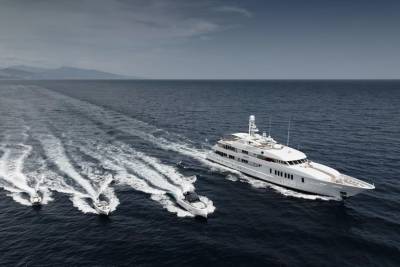 A large white yacht cruises through dark blue water, leaving a wake, with several smaller boats trailing behind it, and a hazy coastline visible in the distance under a cloudy sky.
