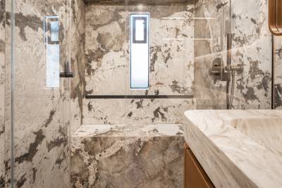 A marble sink in the foreground is set against a shower stall with marble walls and a small window.
