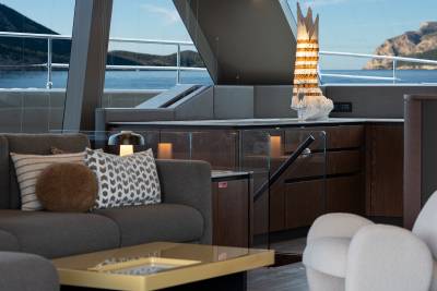 A plush gray sofa with decorative pillows and a golden coffee table are in the foreground, while a modern galley with a unique lamp and a scenic ocean view with mountains are in the background.