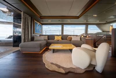 A plush sectional sofa and a unique armchair sit on a textured rug in the foreground, while large windows in the background reveal a cloudy sky and the ocean.