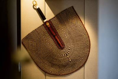 A woven fan with a dark handle and gold loop hangs against a textured, light-colored wall.