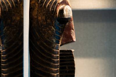 A carved wooden mask with intricate patterns is displayed against a softly lit, blurred background.