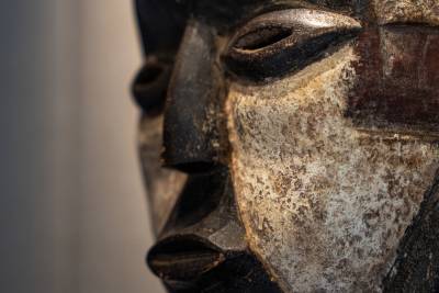 A close-up of a weathered, dark wooden mask with white accents in the foreground is set against a softly blurred, neutral background.